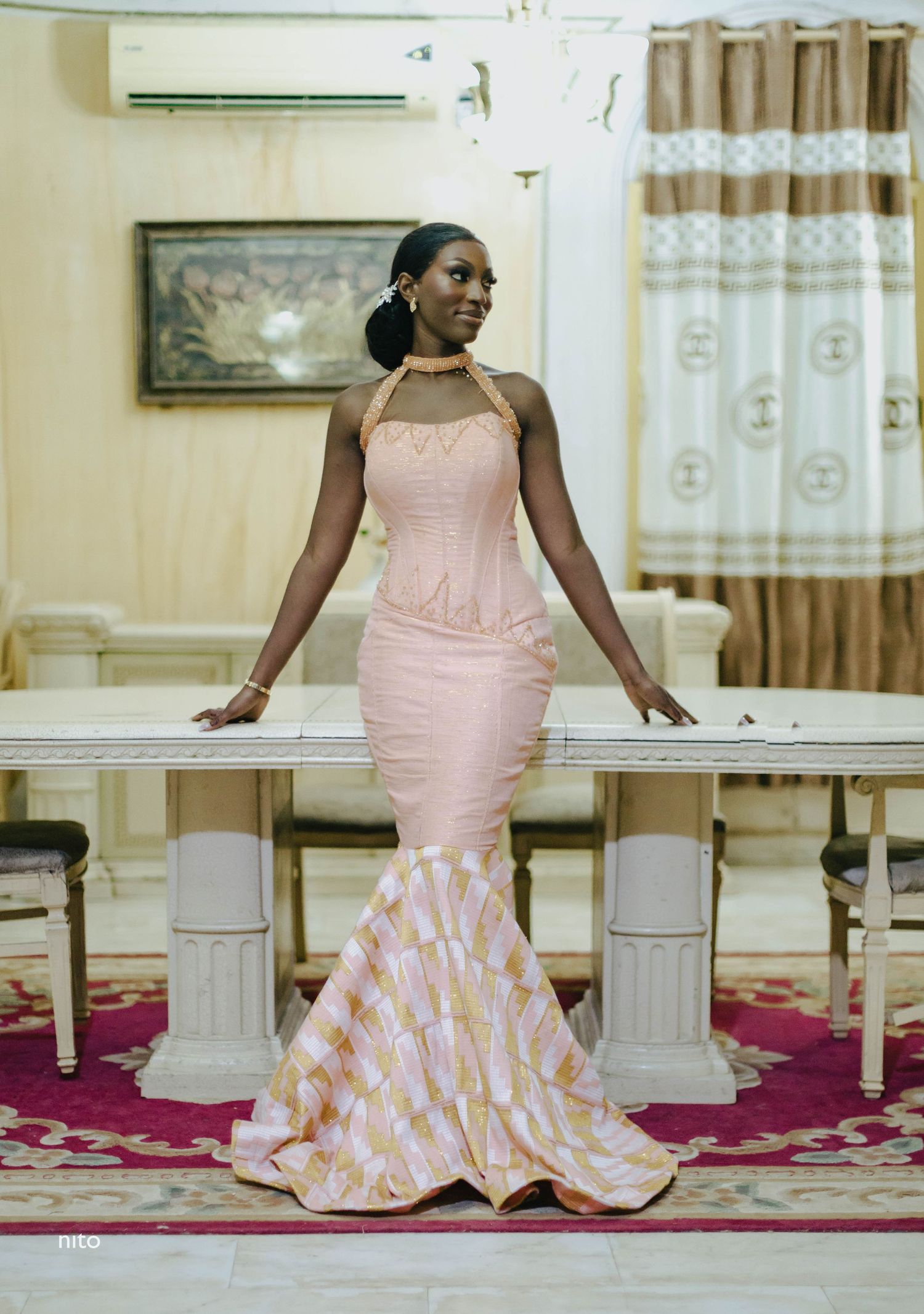 Elegant mermaid-style dress in pink with a patterned flared bottom and halter neckline in an ornate interior setting.