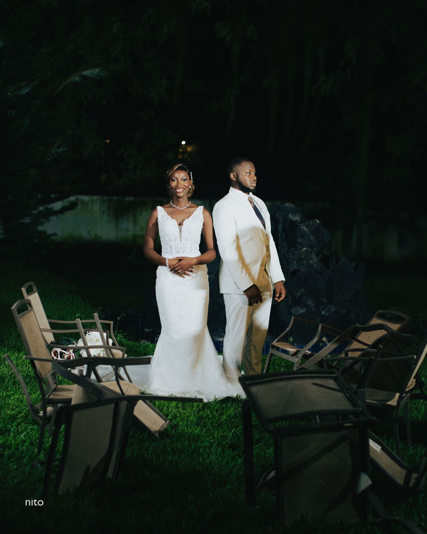 Wedding couple in white attire photographed at night with empty chairs in outdoor setting.