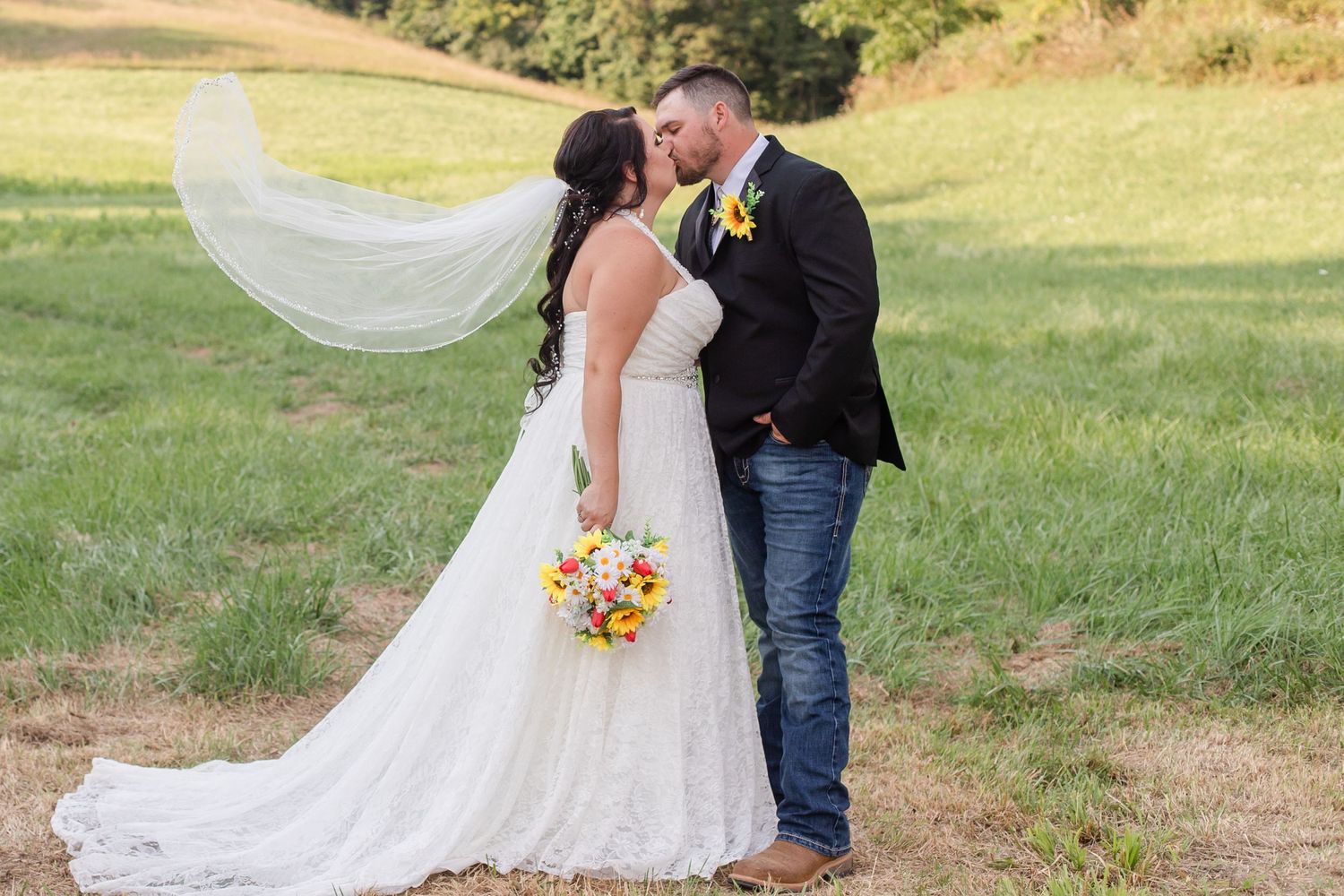 Wedding couple embracing in field with flowing veil and sunflower bouquet.