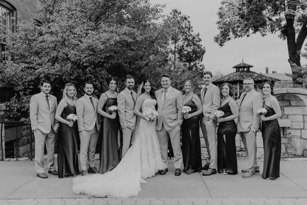 Black and white photo of wedding party posing outdoors with trees and stone building in background.
