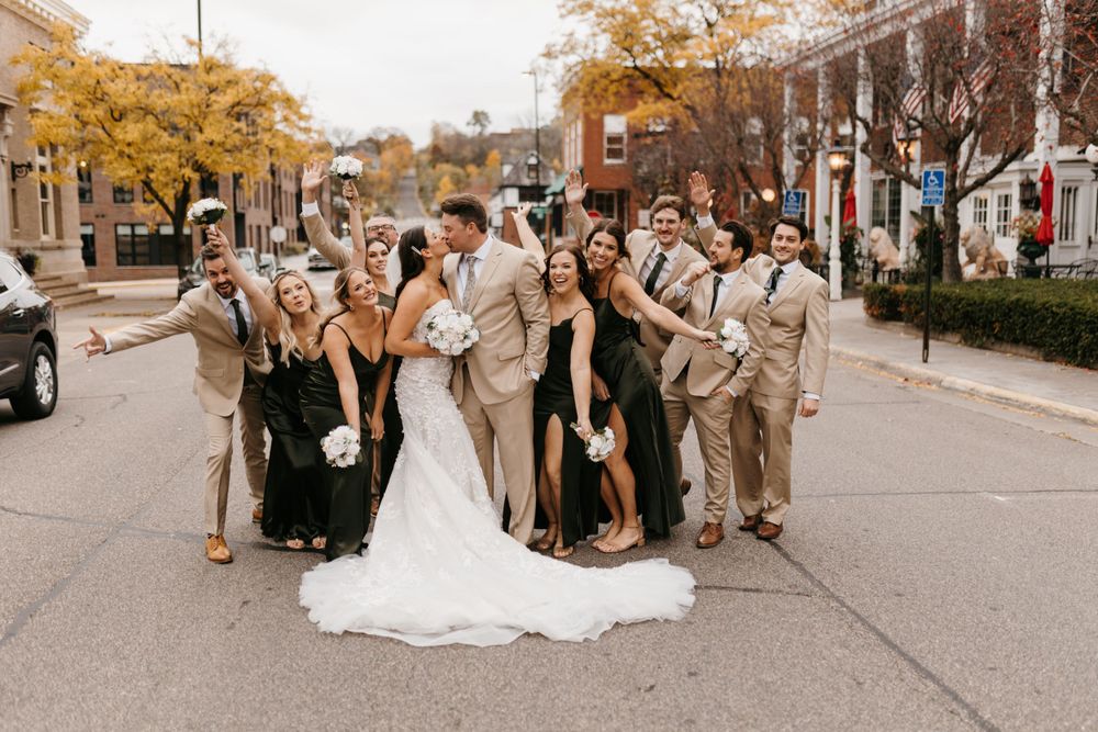 Wedding party surrounds kissing bride and groom on autumn street, celebrating joyfully together.