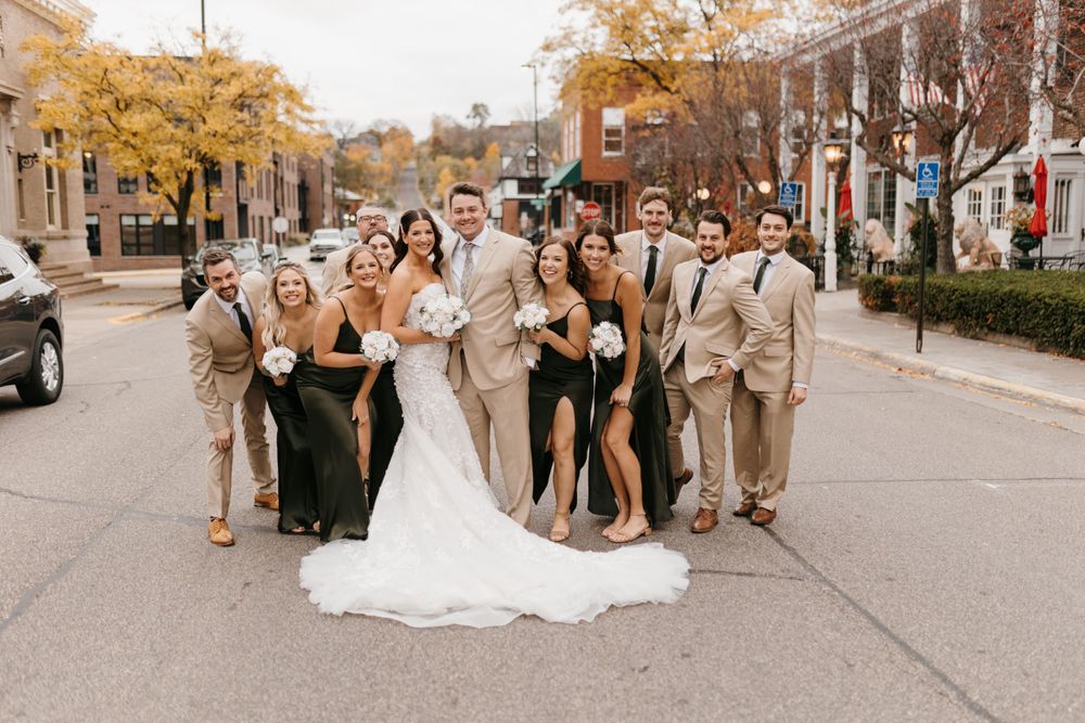Bride, groom and wedding party laugh together on a tree-lined street during fall wedding celebration.