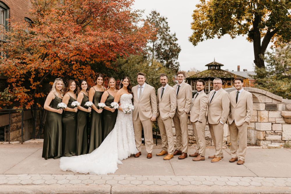 Wedding party lines up outdoors with bridesmaids in olive green gowns and groomsmen in tan suits.