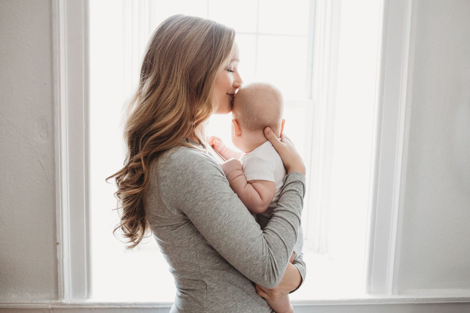 Woman with long curled hair holds her baby close near a window, smiling and pressing her face to his