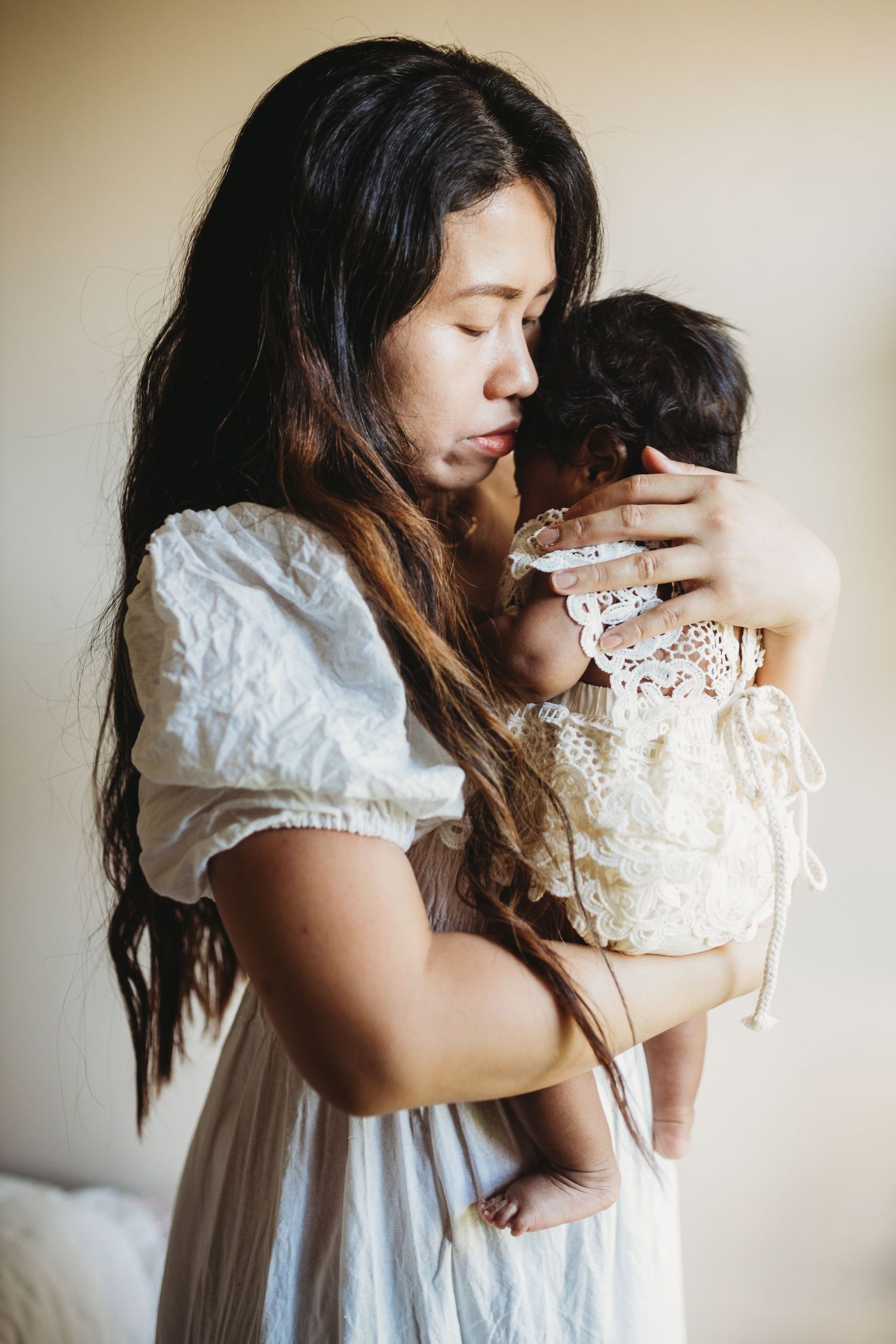 Woman in white dress holding baby wearing lace outfit in soft natural light.