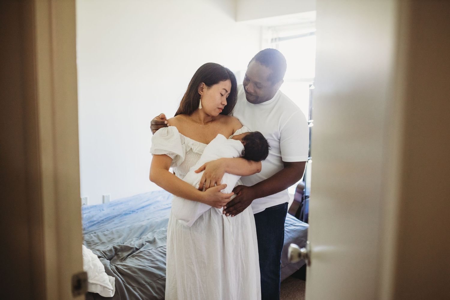Parents in white attire tenderly holding their newborn baby in a sunlit bedroom.