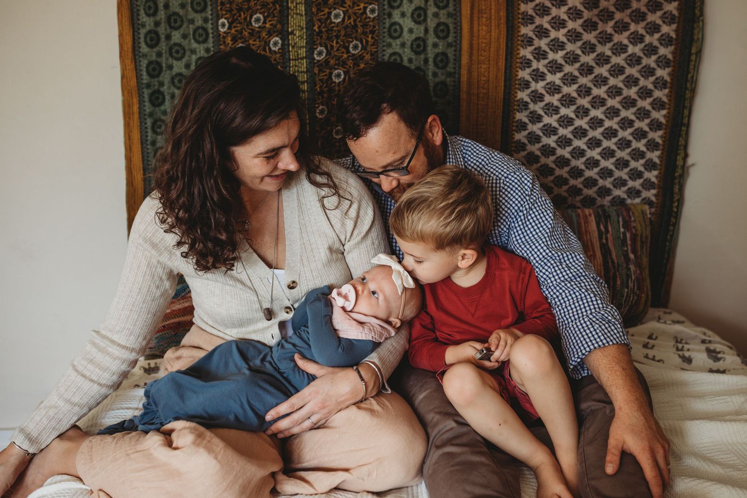 Family and little brother sit on bed admiring infant girl.