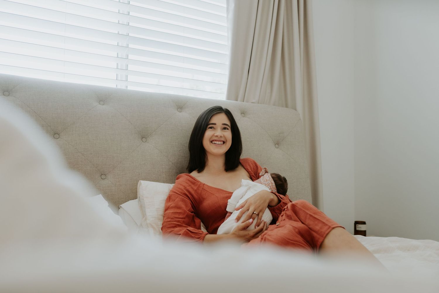 Mother in red dress relaxes on grey couch while holding newborn.