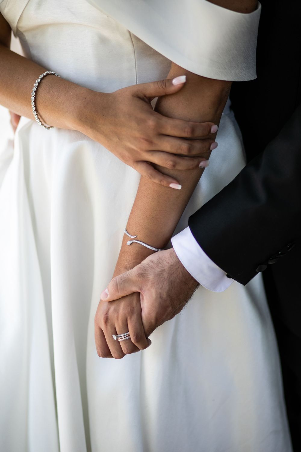 Close-up of hands with wedding rings and jewelry against a white wedding dress.