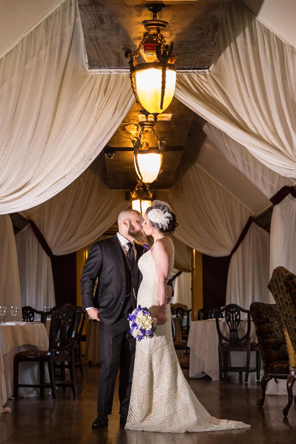 Elegant wedding couple embraces under draped ceiling with vintage lanterns casting warm glow in ballroom.