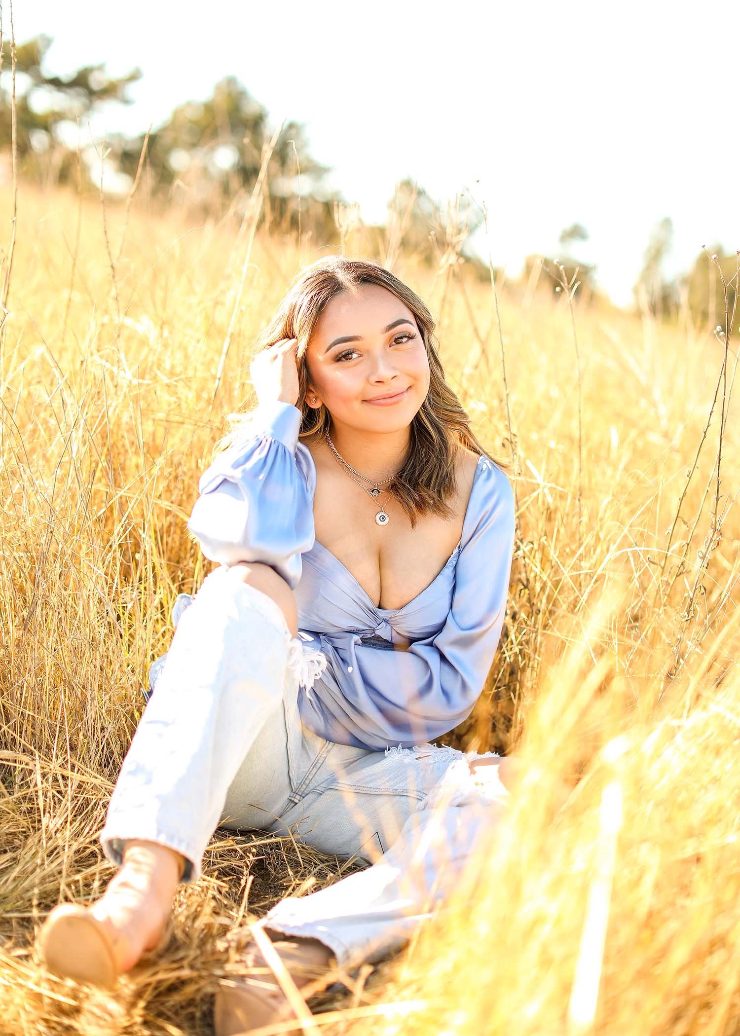 Woman in blue outfit sitting in tall golden grass during sunset portrait session.