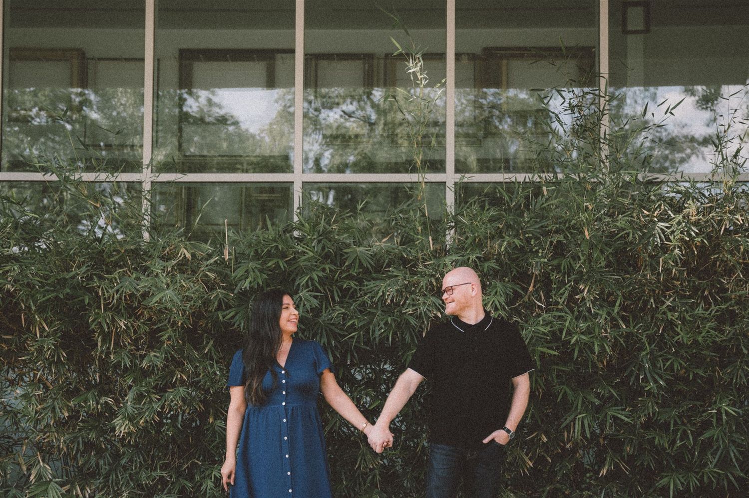 A couple holding hands in casual attire stand against a wall with greenery and windows.
