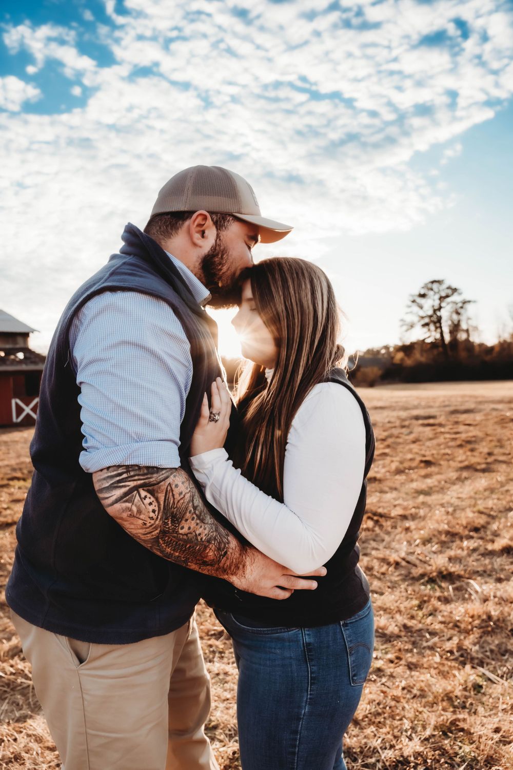 Couple sharing a tender kiss at golden hour in a farm field, man with tattoo sleeve wearing a cap. Jacksonville, NC