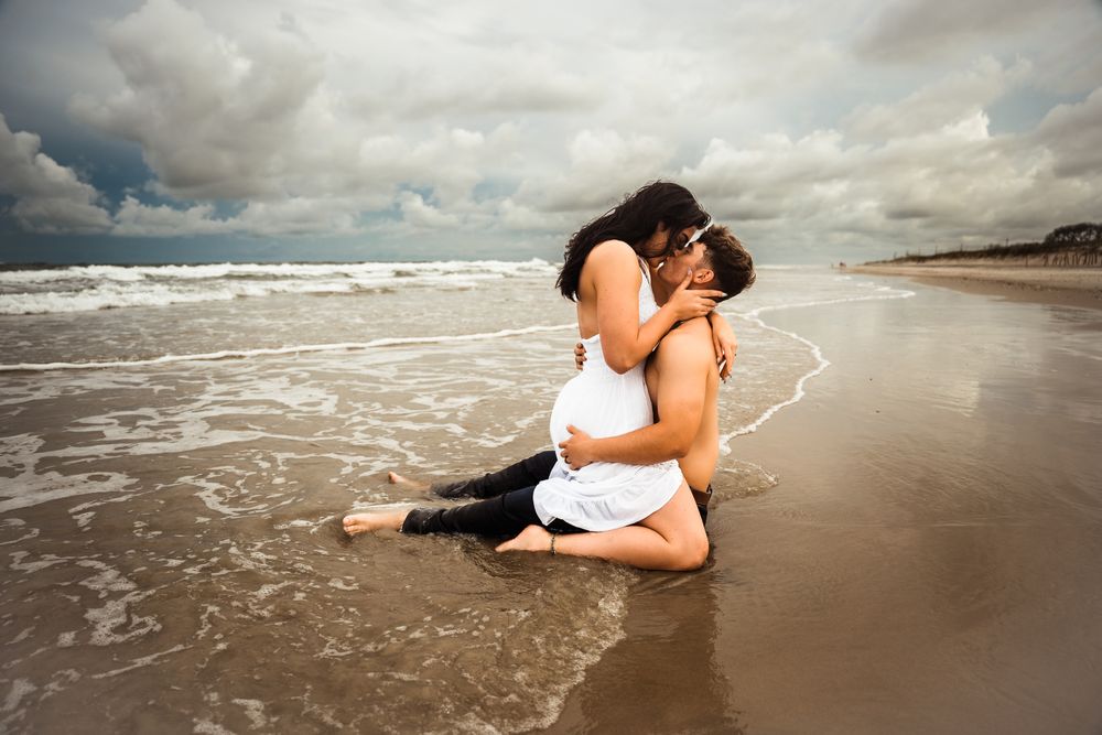 Couple sharing a romantic kiss while sitting in ocean waves on a sandy beach under cloudy skies. Jacksonville, NC