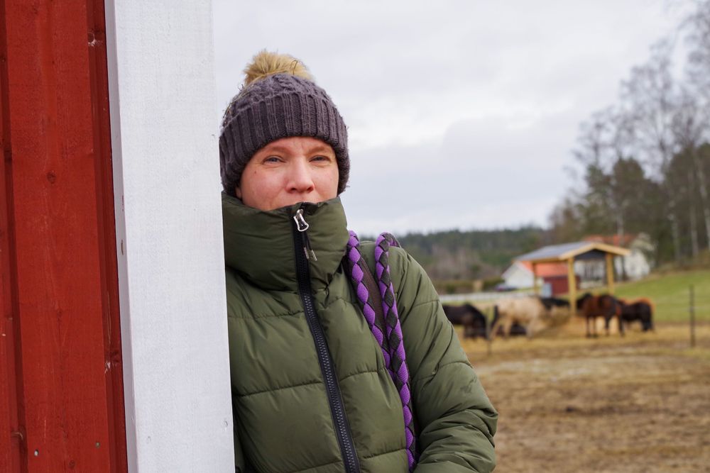 Someone in a puffy green winter coat and knit hat stands against a red wall with farm buildings behind.