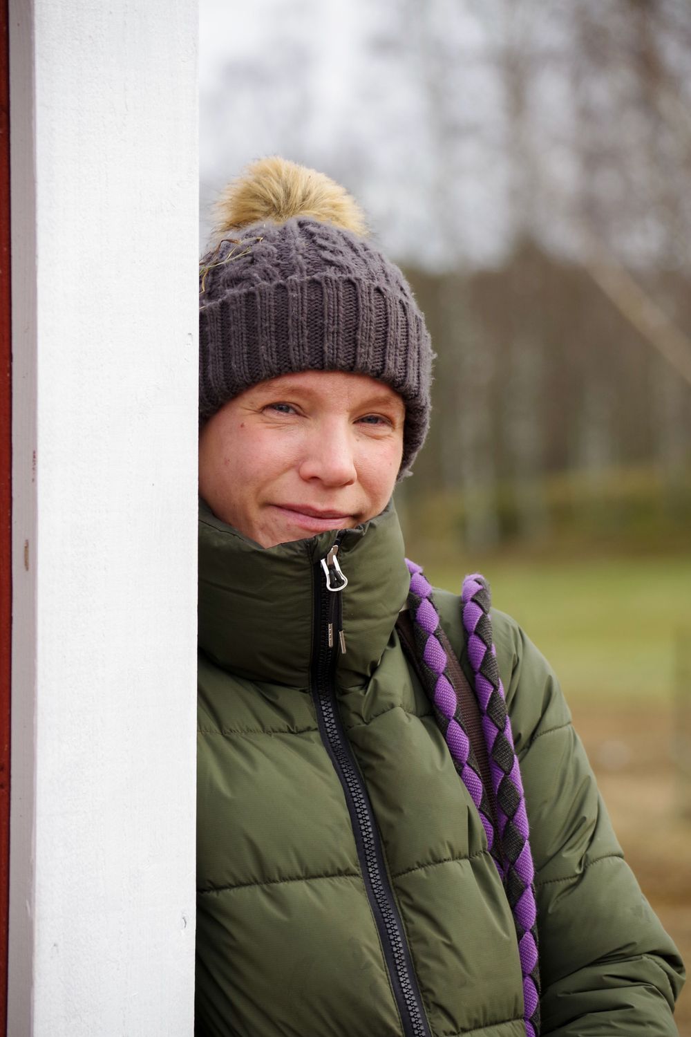 Person wearing a green winter coat and gray knit beanie with pom pom stands outdoors in cold weather.