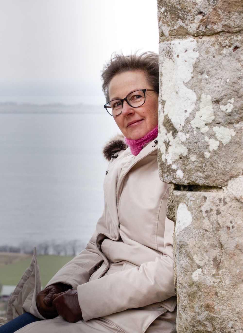 Figure in beige coat and glasses peeks around stone wall corner with misty landscape in background.