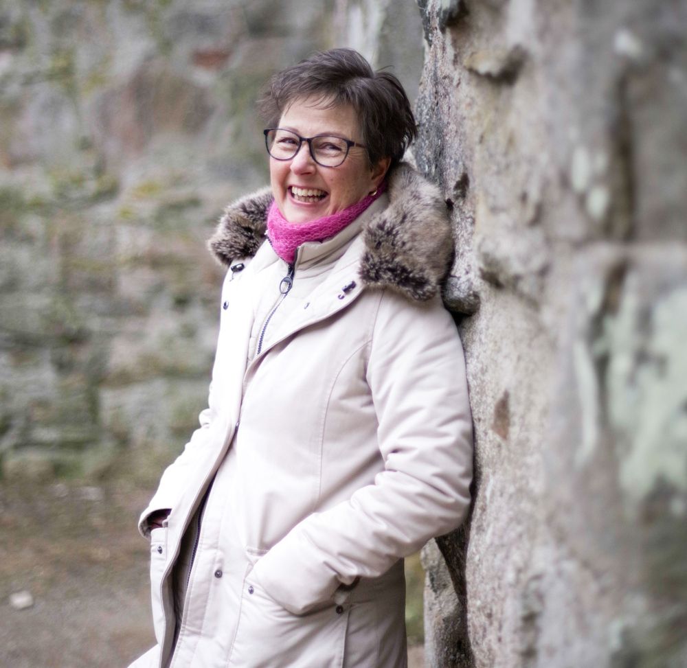 Person in cream winter coat and pink scarf laughs joyfully while leaning against stone wall outdoors.
