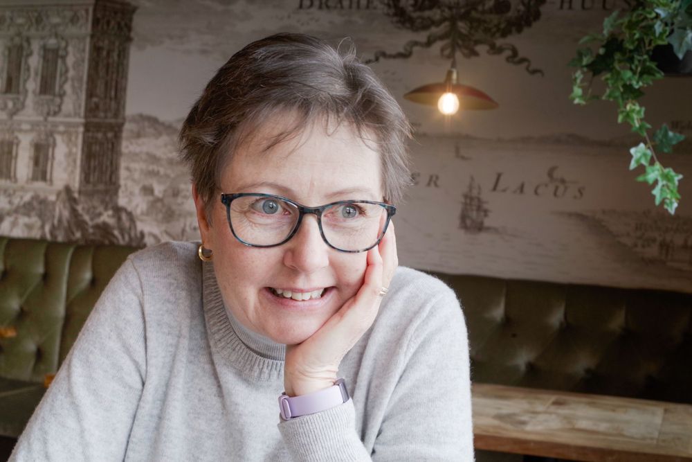 Person in gray sweater and glasses smiles warmly while resting in a cozy indoor setting.