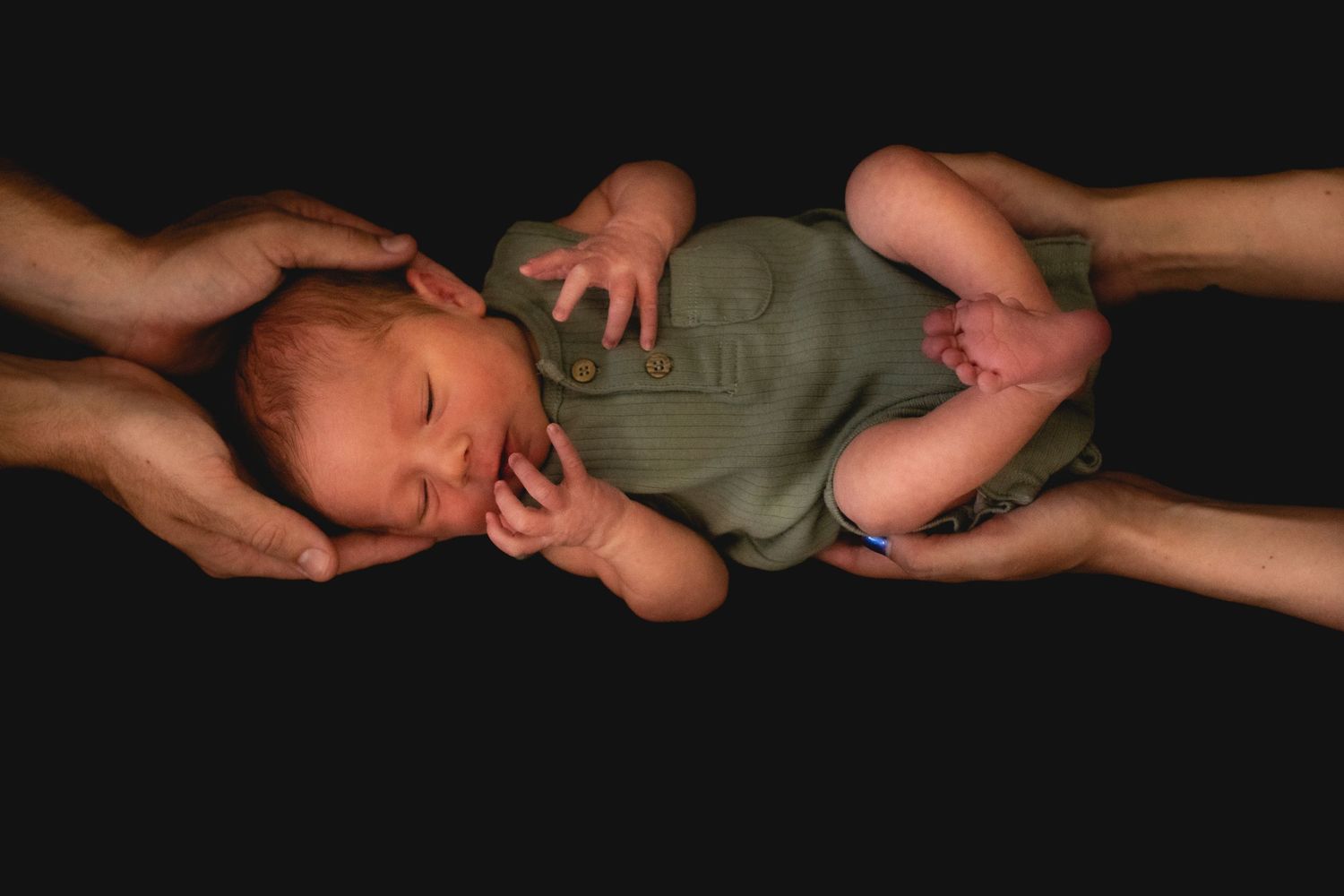 Newborn baby supported by adult hands against dark background.
