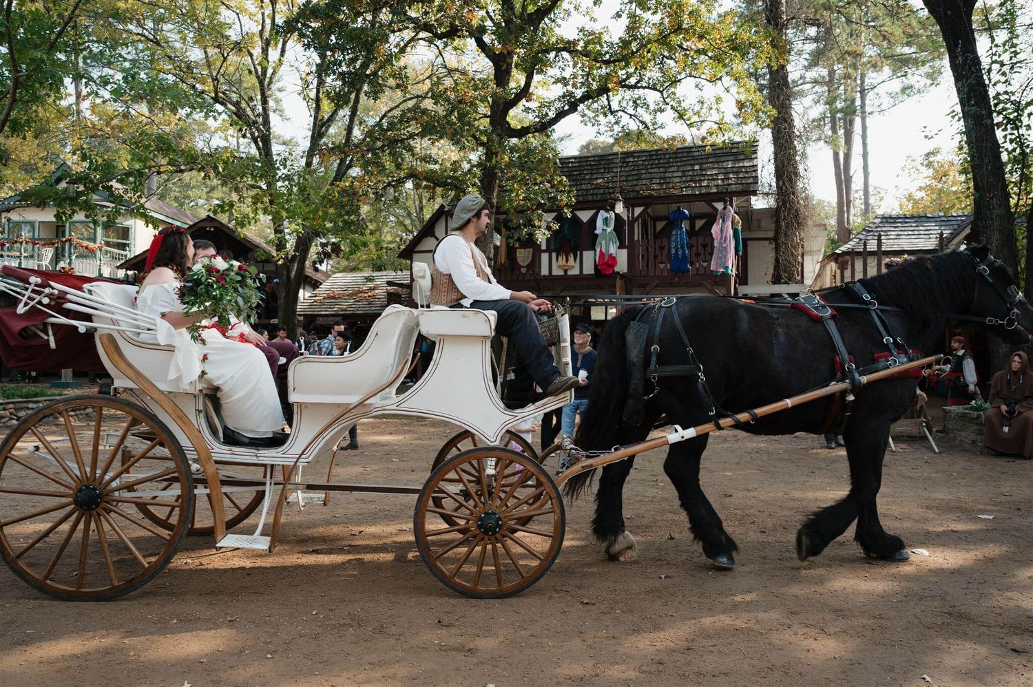 A pair of black horses pull an elegant white carriage through a renaissance faire grounds.