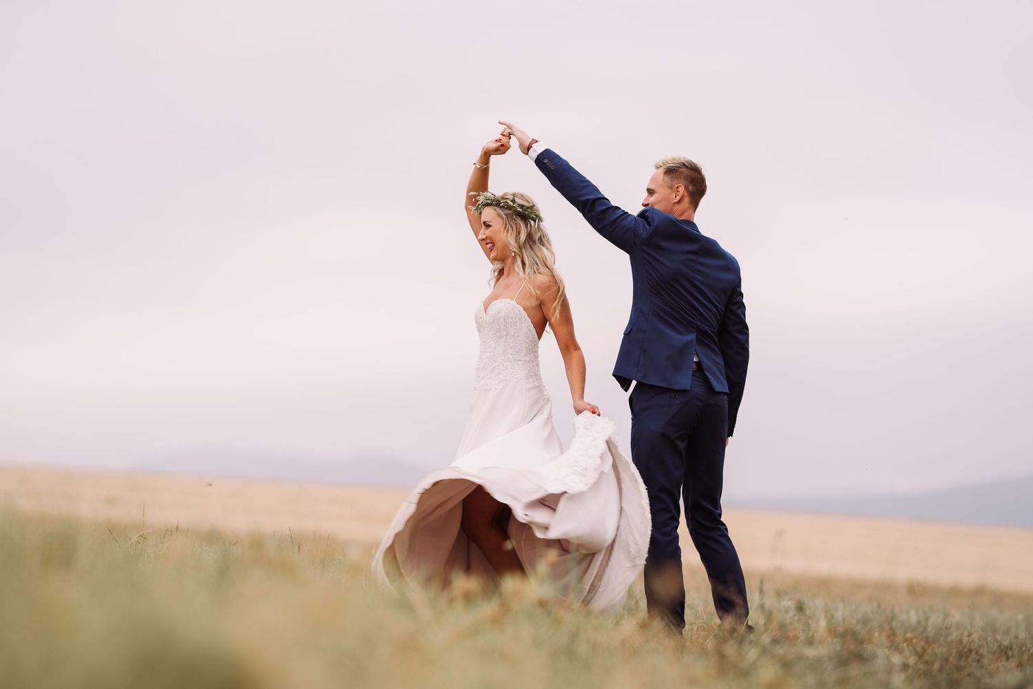 Groom twirling bride in a flowing dress in an open golden field under a soft cloudy sky.