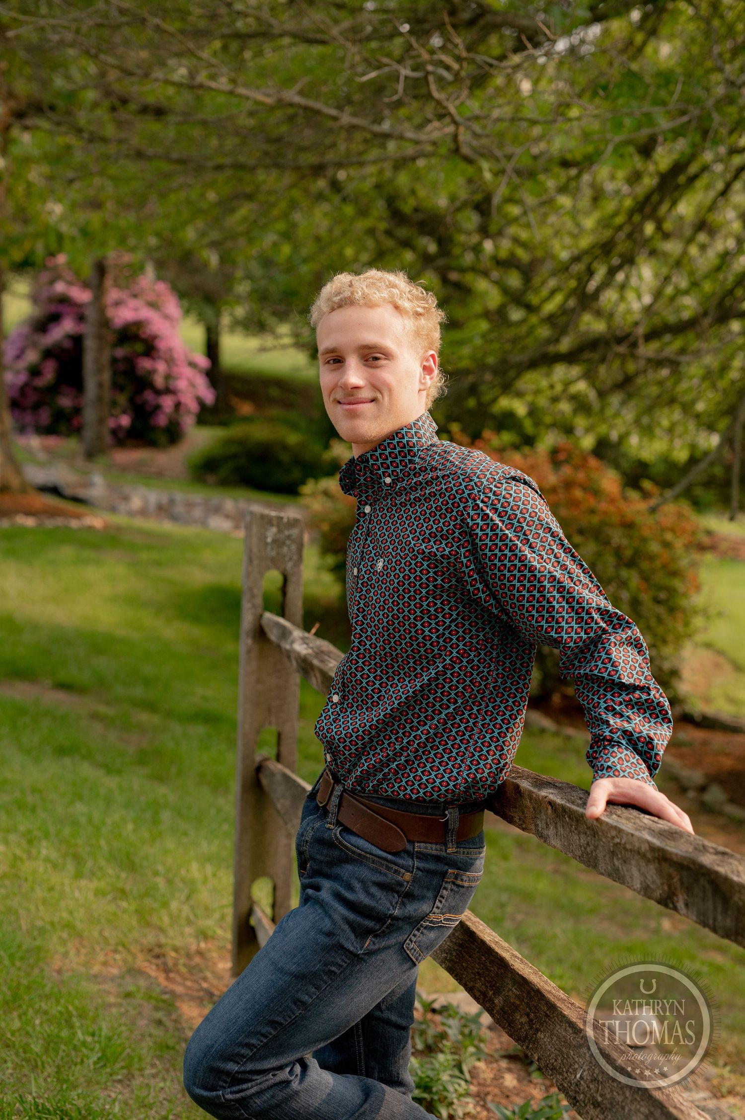 Casual outdoor portrait by wooden fence with flowering bushes and trees in background.