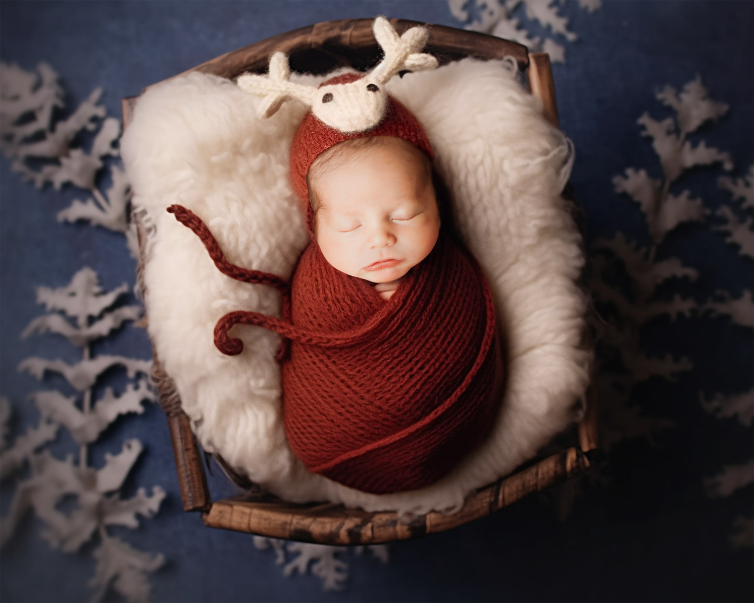 A cozy newborn photography setup features a rustic wooden bowl with white fluff and burgundy wrap against a snowy background.