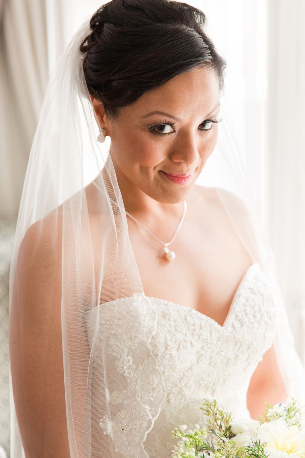 A bride in a white lace wedding dress holding a bouquet looks down with a gentle smile.