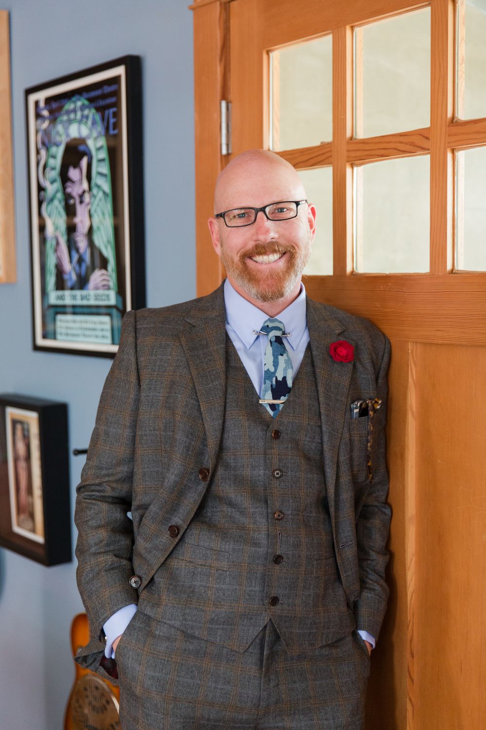 A groom in a grey plaid suit with a red boutonniere stands near wooden window frames.