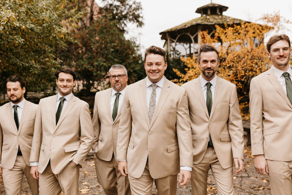 Groomsmen in tan suits walking confidently outdoors surrounded by golden autumn trees.