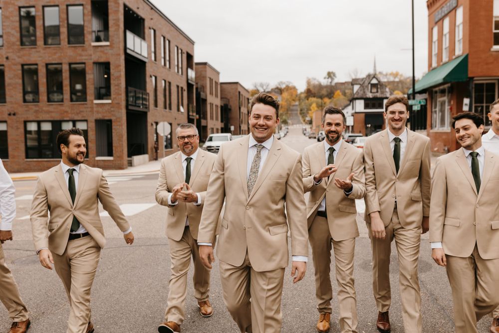 Groom leads groomsmen in tan suits laughing while walking through city streets on wedding day.