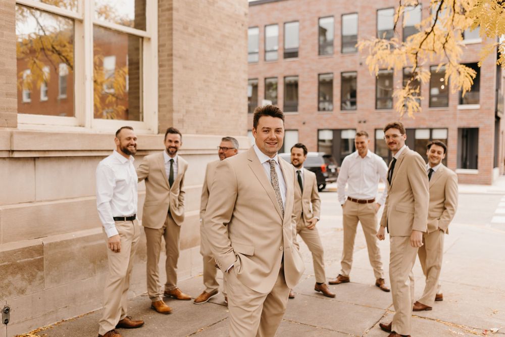 Groom poses in tan suit with groomsmen in white shirts gathered behind near brick building.