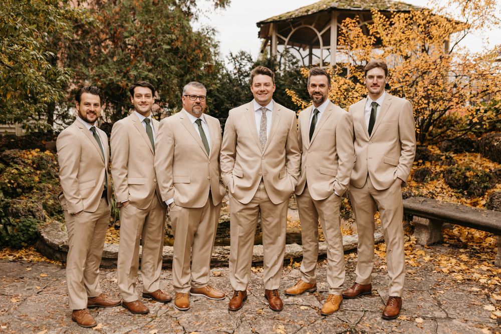 Six groomsmen in matching tan suits and dark ties smiling outdoors with fall leaves behind them.