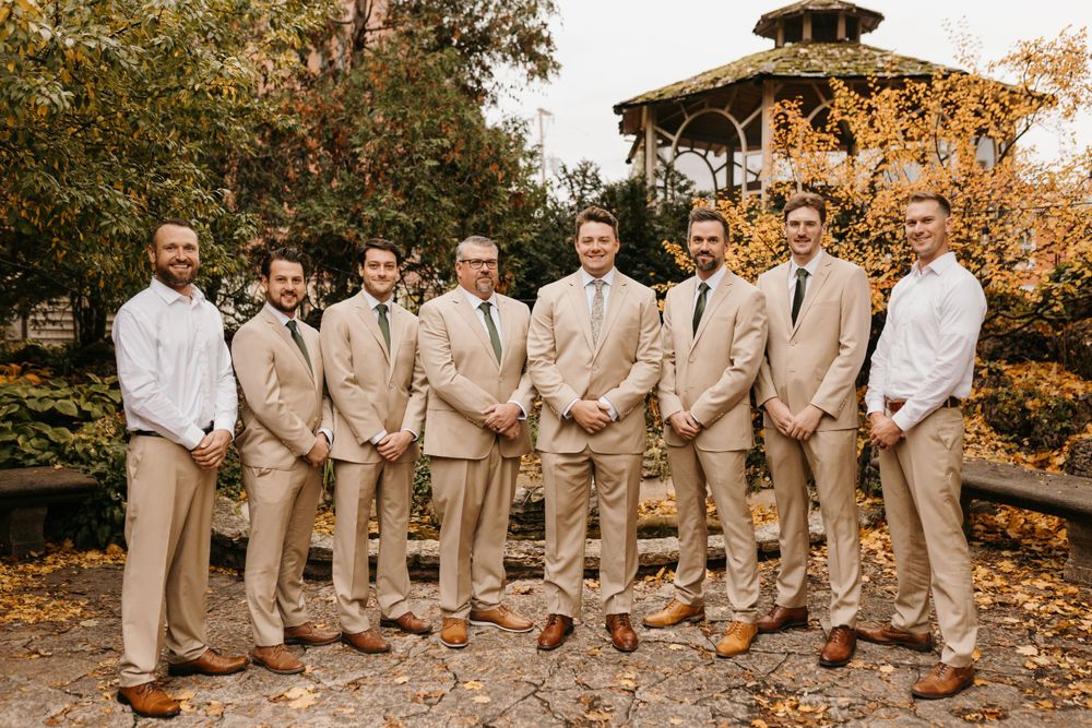 Groom and groomsmen in tan suits posing outdoors near gazebo with autumn foliage backdrop.