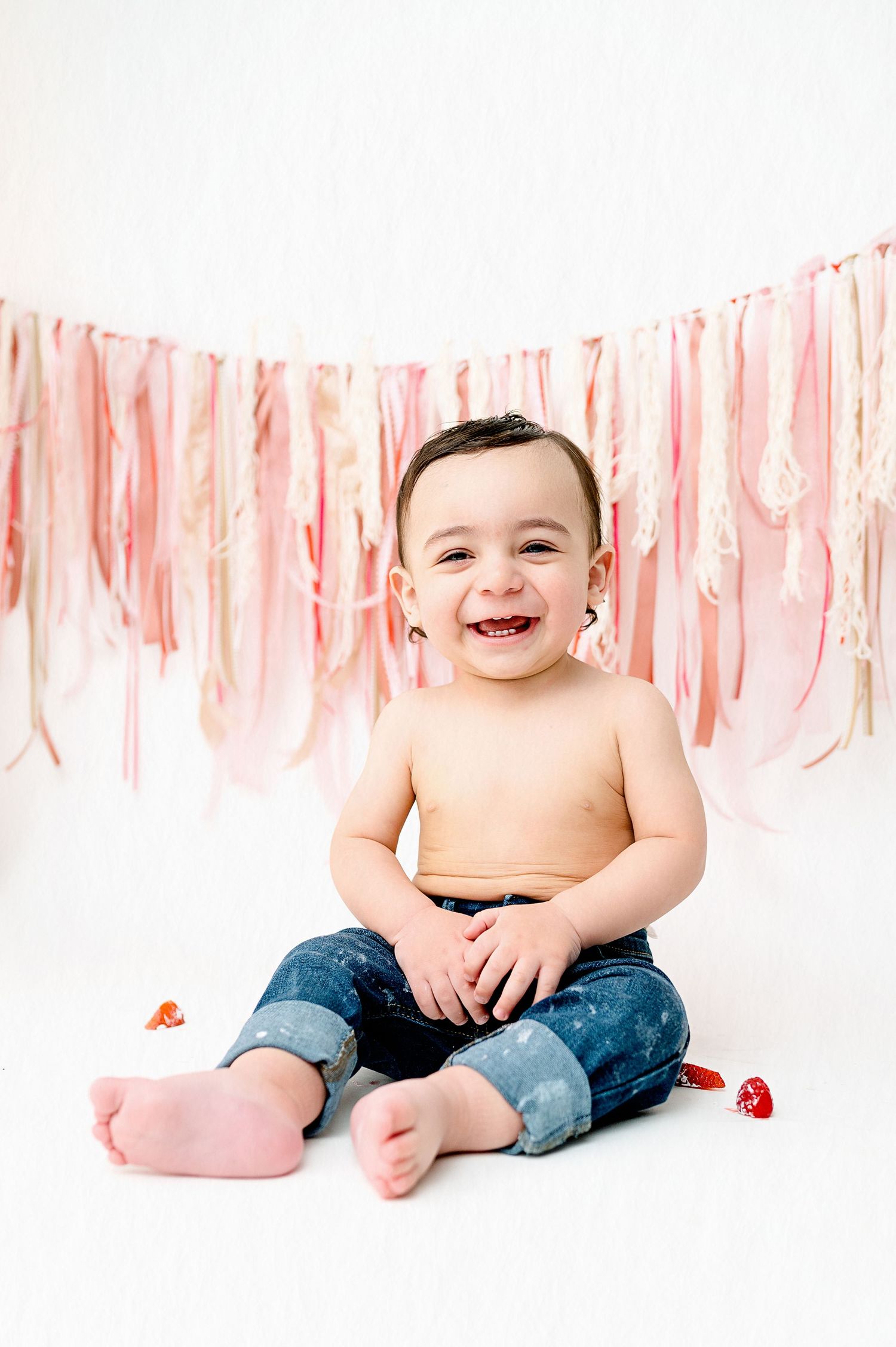 Happy baby in jeans sits on white floor with pink party streamers and red hearts.