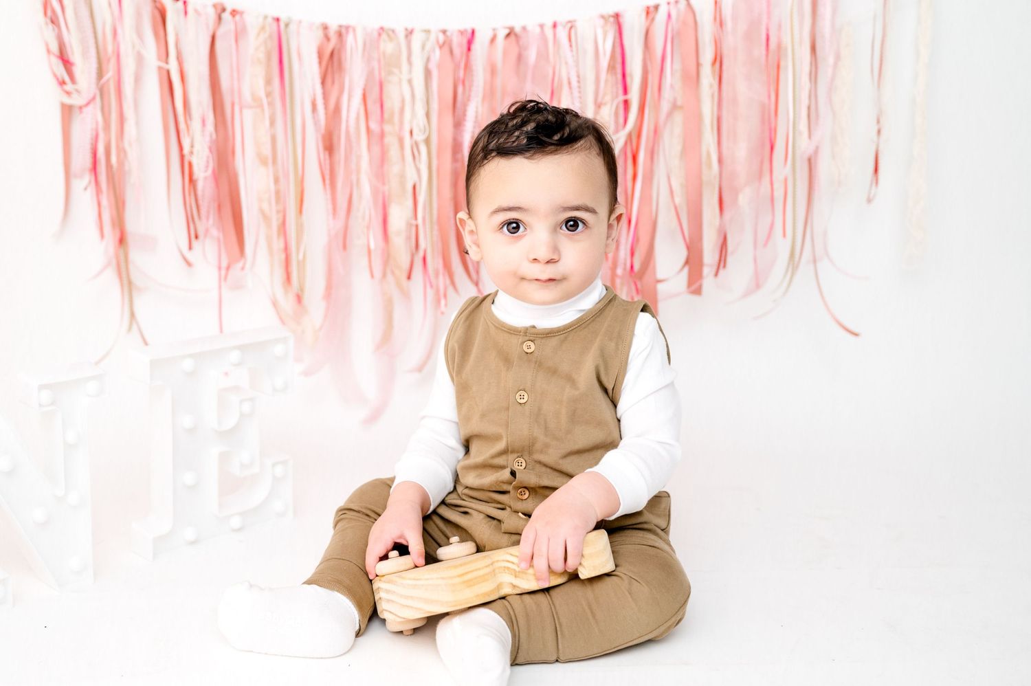 Smiling baby in brown outfit sits against white background with pink tissue paper garland.