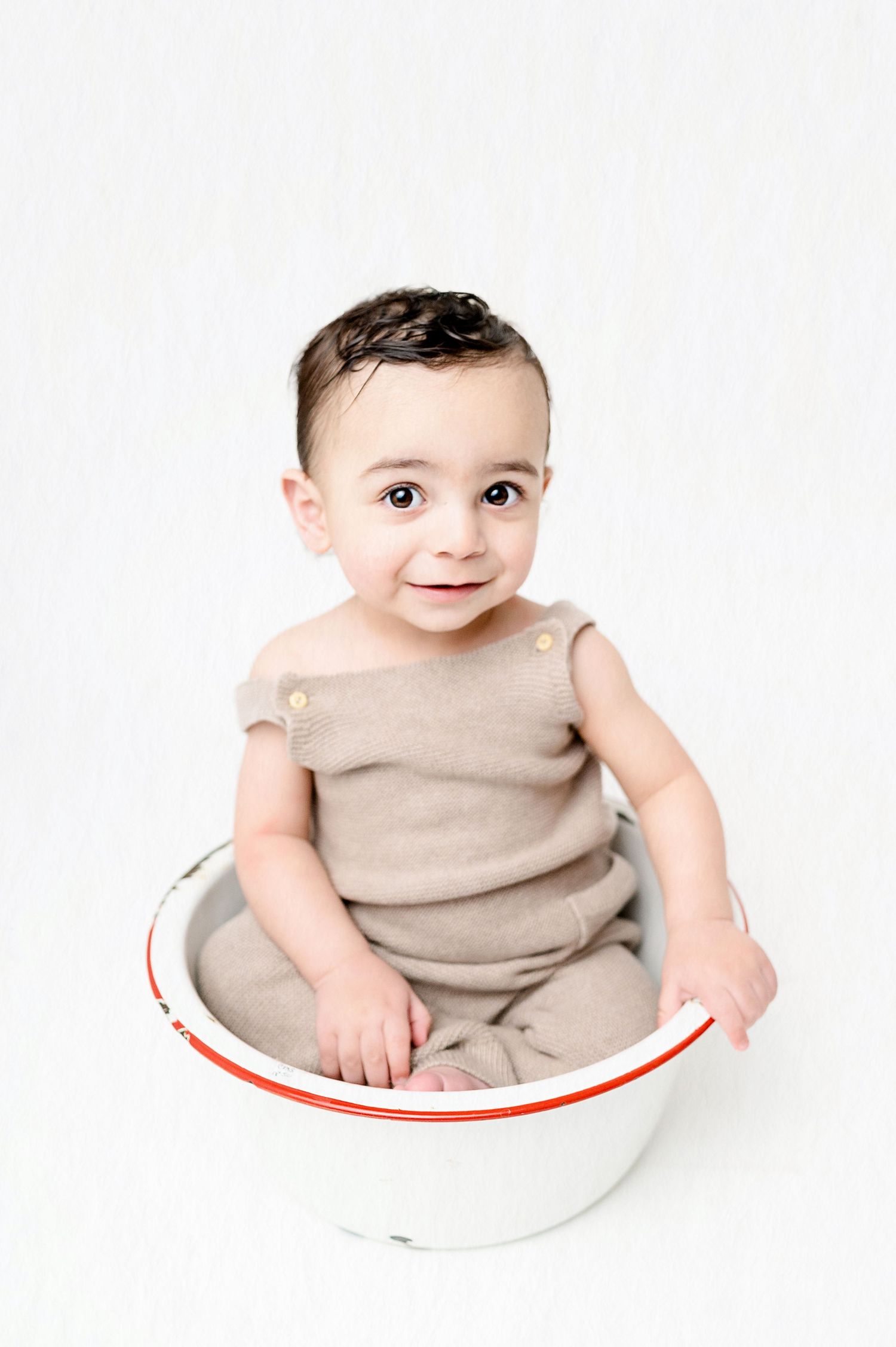 Baby in beige sleeveless outfit sits inside white bowl with red trim.