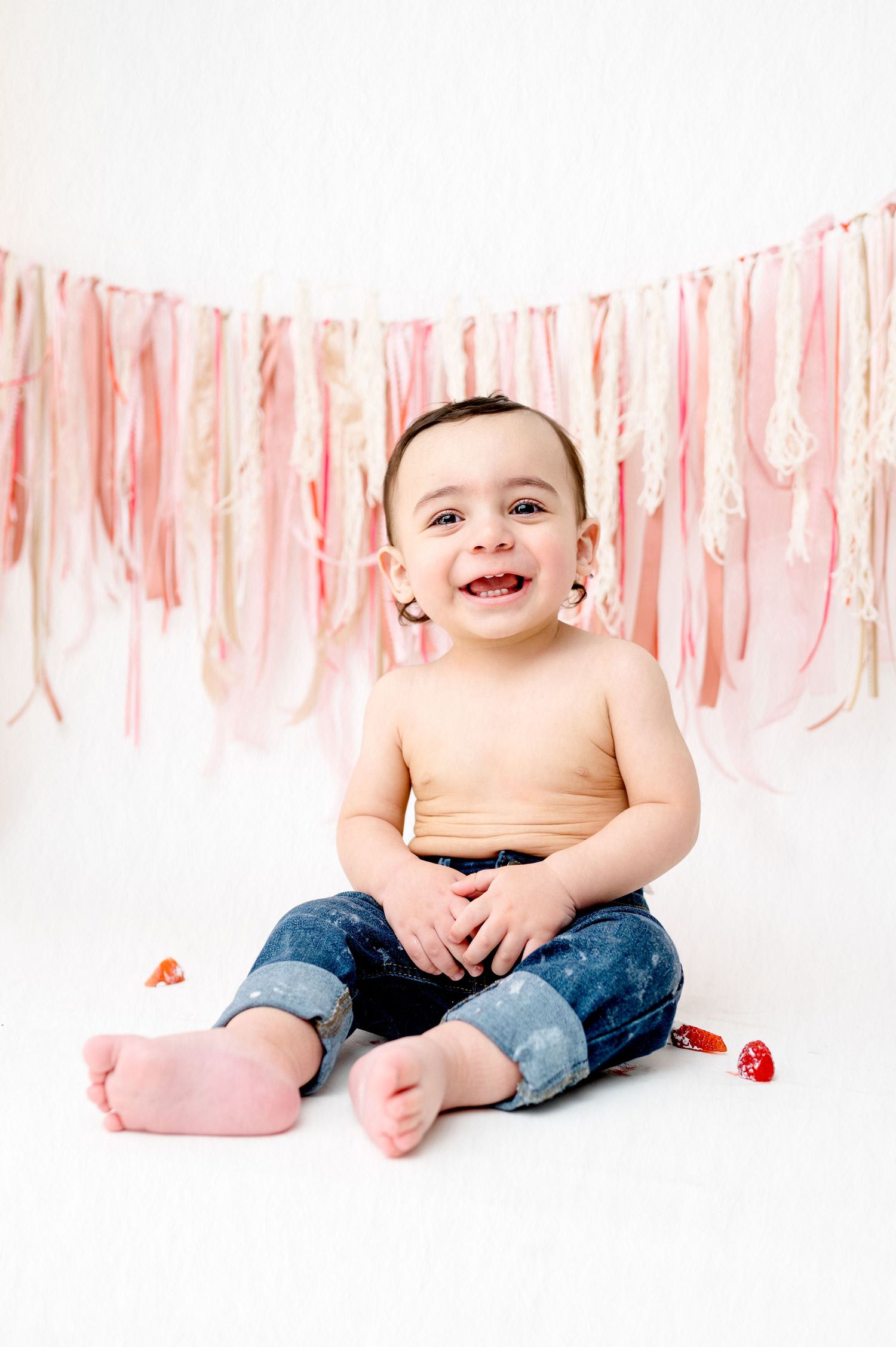 Happy baby in jeans sitting against pink streamer backdrop.