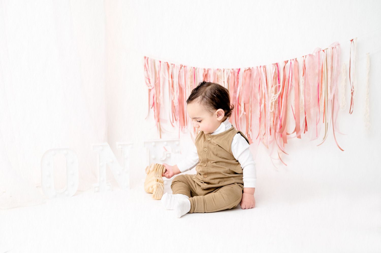 Infant wearing brown romper plays on white backdrop under pink party decorations.