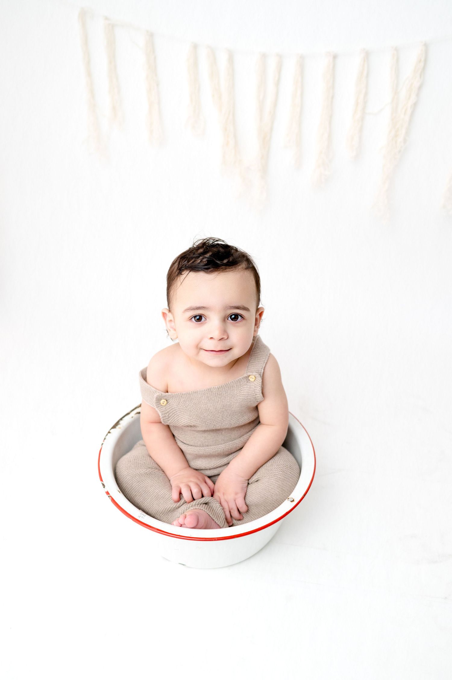 Baby in beige outfit sitting in white enamel bowl against light background.