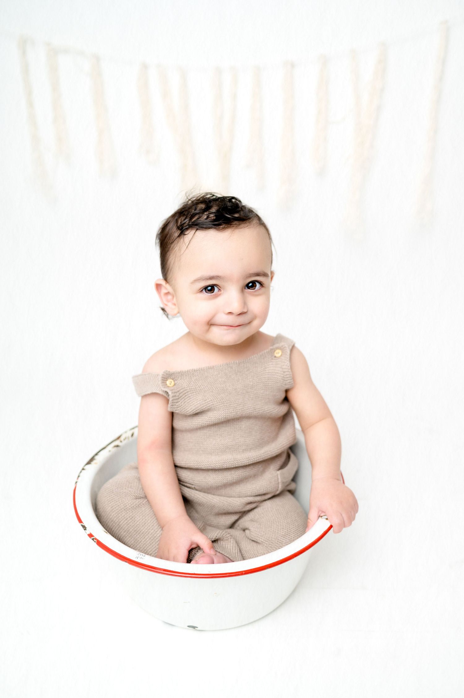 Infant wearing beige outfit smiles while sitting in white bowl with red trim.