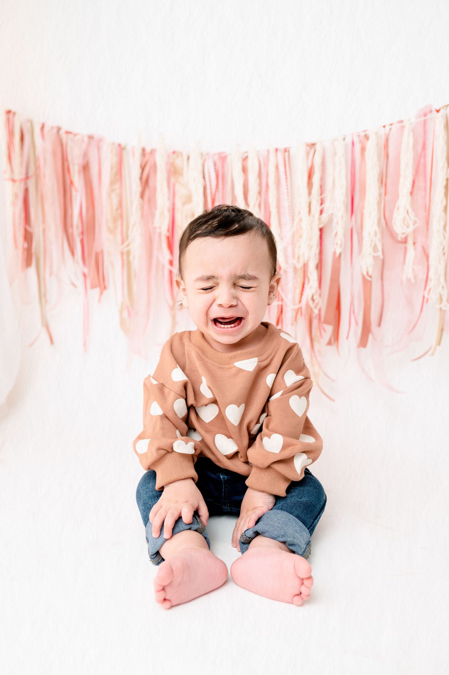 Crying baby wearing heart patterned sweater sitting against pink streamer backdrop.