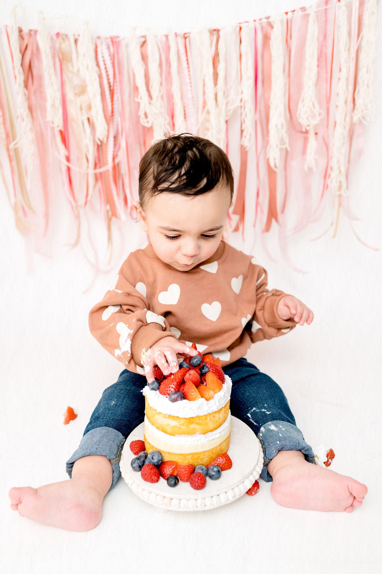 Baby exploring birthday cake decorated with fresh berries against pink streamer wall.