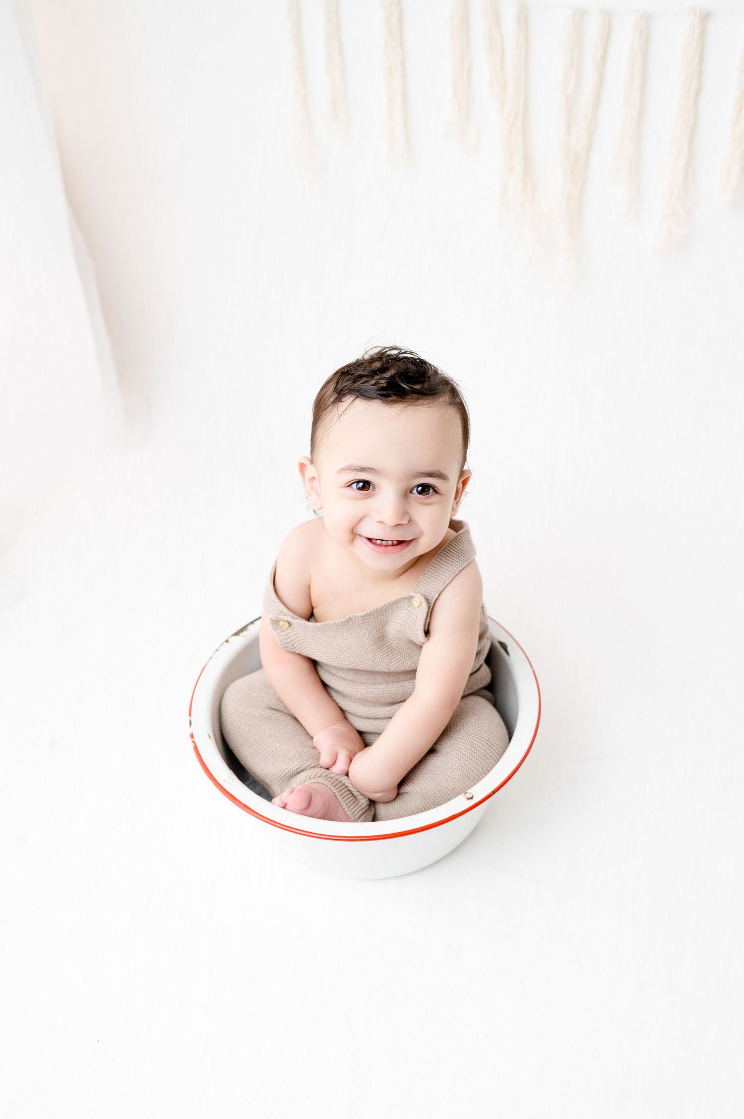 Smiling baby wearing tan romper seated in white enamel bowl with decorative backdrop.