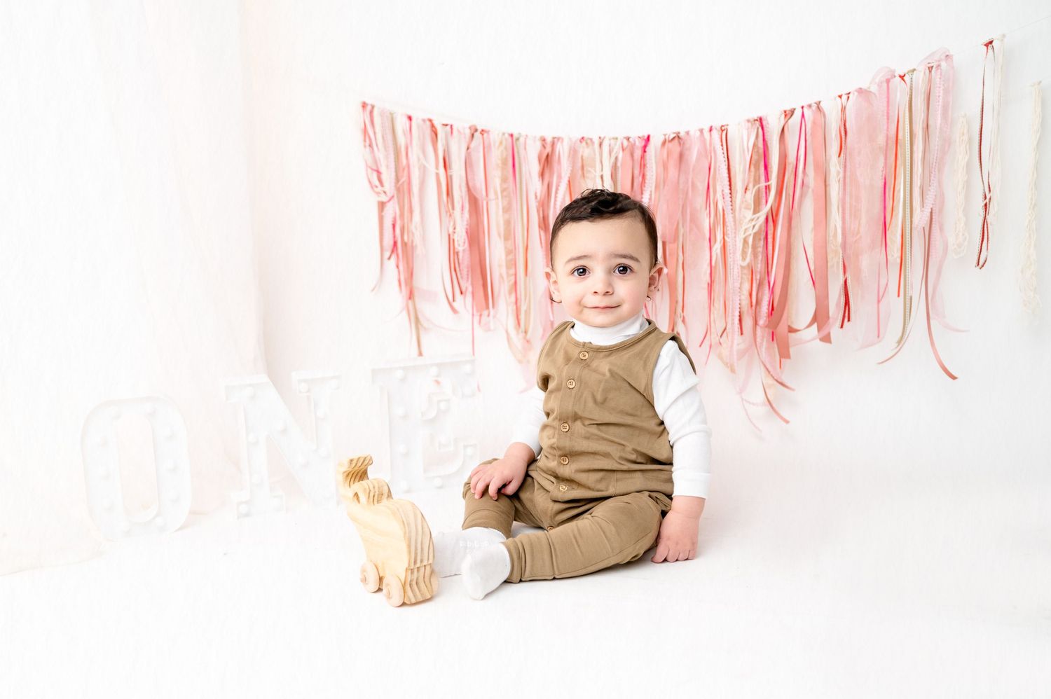 Baby in brown outfit sits on white floor with pink streamers hanging in background.