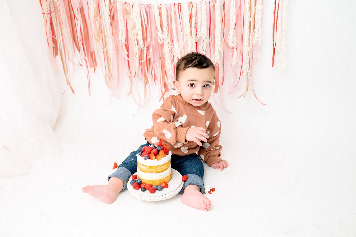Happy baby in heart sweater with birthday cake and pink streamers.