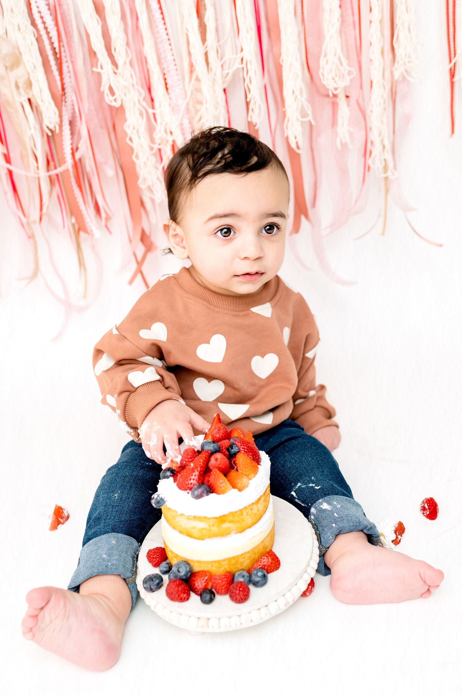 Baby in brown heart sweater examining fruit-topped cake against pink backdrop.