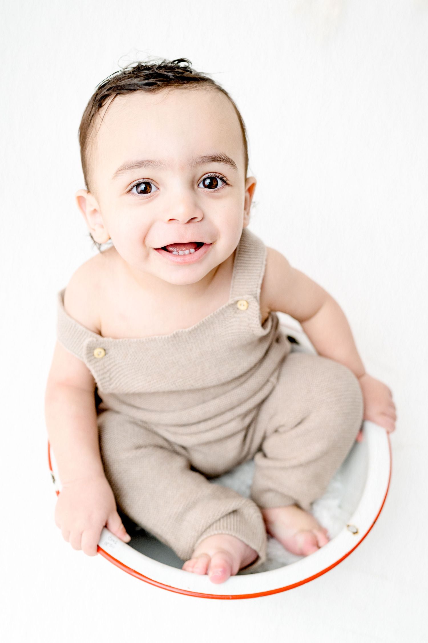 Infant smiles while sitting in white and red rimmed bowl wearing tan outfit.