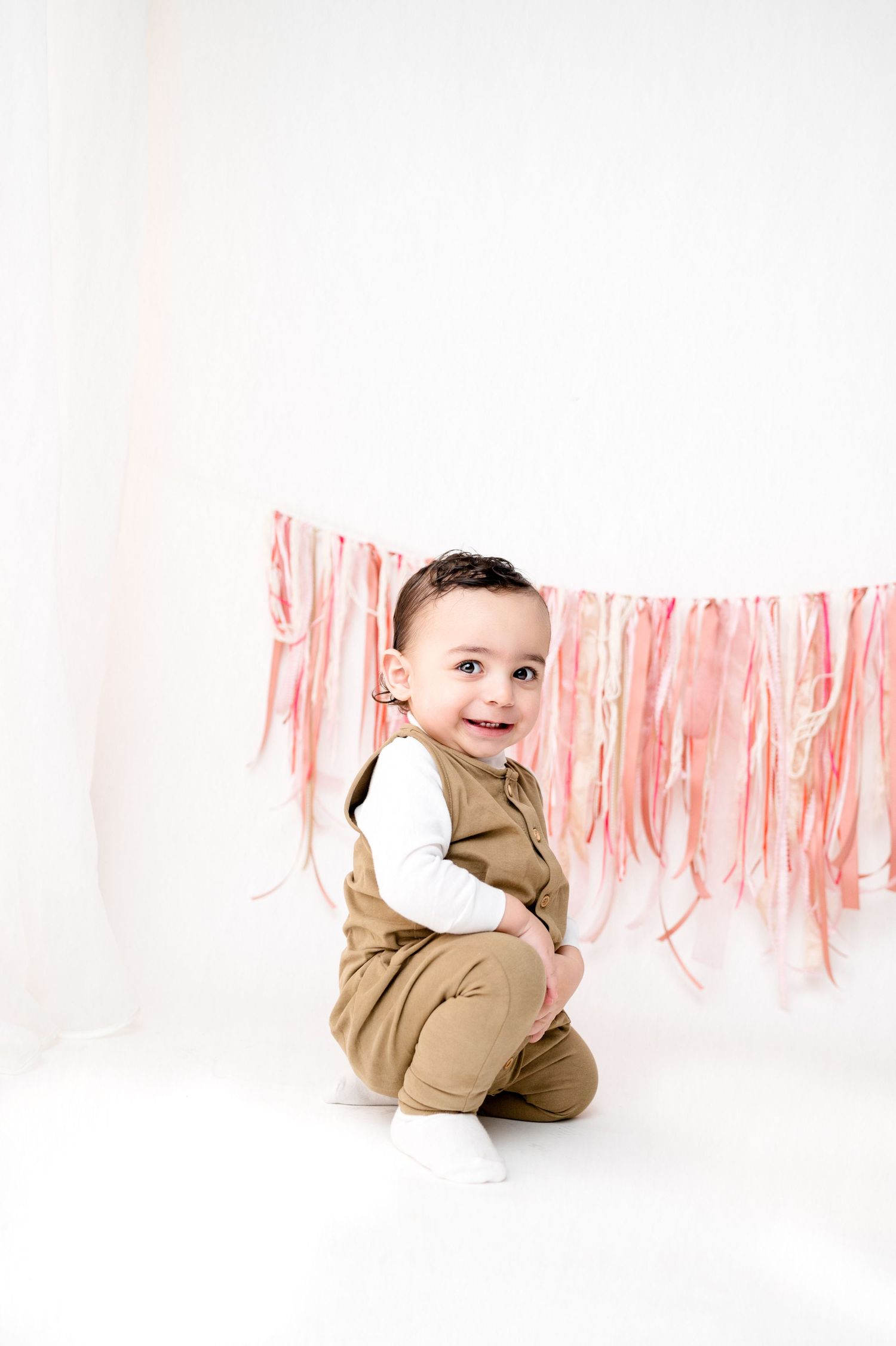 Cheerful infant poses on white floor with pink streamer decorations overhead.